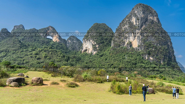 Picturesque karst mountains against a clear blue sky. There is a round hole at the top. Green vegetation on steep slopes, in the valley. Boulders in the meadow. People are walking around the clearing. Стоковое фото, фотограф Вера Ивановна Кубарева / Фотобанк Лори
