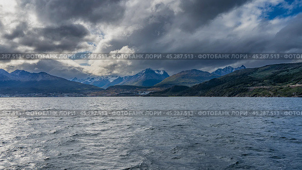 Picturesque snow-capped Andes mountains against a blue sky, clouds. The city of Ushuaia is on the ocean. The water surface is in the foreground. The Beagle Channel. Argentina. Tierra del Fuego (2023 год). Стоковое фото, фотограф Вера Ивановна Кубарева / Фотобанк Лори