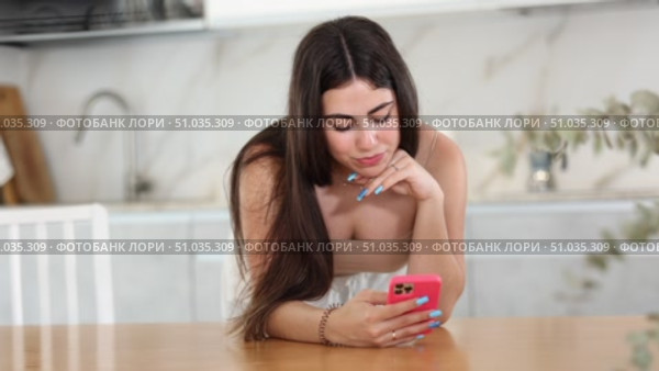 Pleased young woman leaning on table and watching something interesting on her mobile phone in the kitchen. Стоковое видео, видеограф Яков Филимонов / Фотобанк Лори