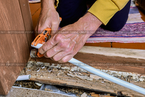 Plumber uses hand pipe cutter to cut PEX pipe near wooden floor, preparing pipe end for connection during plumbing work in residential building. Стоковое фото, фотограф Владимир Григорьев / Фотобанк Лори