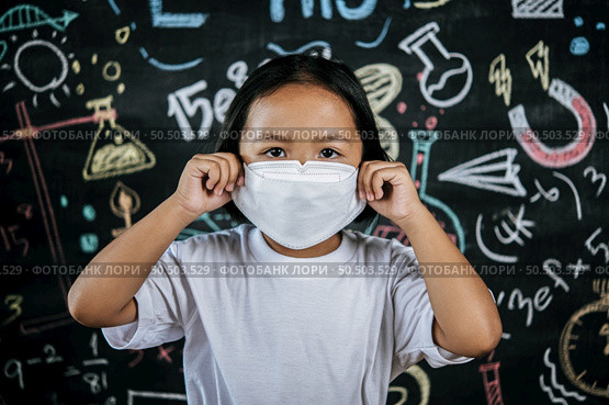 Portrait of a little girl wearing eyeglasses and facemask at school to avoid the spread of coronavirus, new normal with schoolchild,  blure blackboard in background. Стоковое фото, агентство Ingram Publishing / Фотобанк Лори