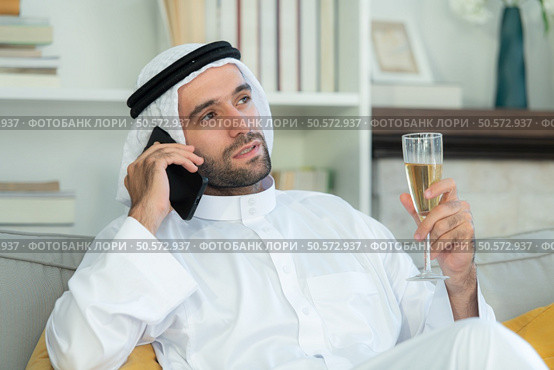 Portrait of a smiling Arabian man holding a glass of champagne and using mobile phone at home. Стоковое фото, агентство Ingram Publishing / Фотобанк Лори