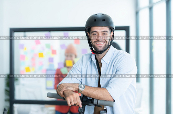 Portrait of smiling businessman riding electric scooter in office with colleague in background. Стоковое фото, агентство Ingram Publishing / Фотобанк Лори