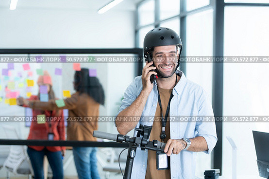 Portrait of smiling businessman talking on mobile phone while colleague using scooter in office. Стоковое фото, агентство Ingram Publishing / Фотобанк Лори