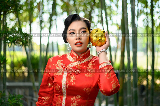 Portrait pretty Asian woman in a Chinese cheongsam posing with piggy bank on her cheek in bamboo forest background, copy space, Chinese New Year concept. Стоковое фото, агентство Ingram Publishing / Фотобанк Лори
