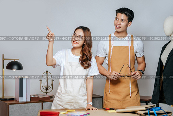 Portrait Young handsome man and pretty woman in glasses wearing apron, Couple fashion designer working together with happy, mannequin and tailoring tools on desk at studio. Стоковое фото, агентство Ingram Publishing / Фотобанк Лори