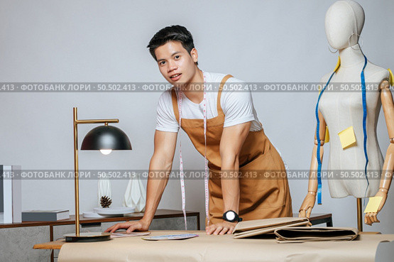 Portrait Young male fashion designer working with fabric and clothing sketches at the studio at home, full of tailoring tools, model and equipment on table. Стоковое фото, агентство Ingram Publishing / Фотобанк Лори