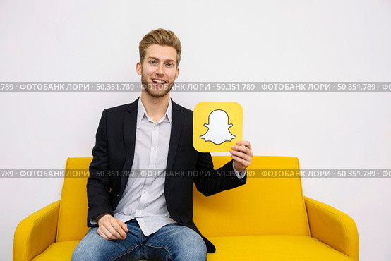portrait young man sitting yellow sofa holding snapchat icon. Стоковое фото, агентство Ingram Publishing / Фотобанк Лори