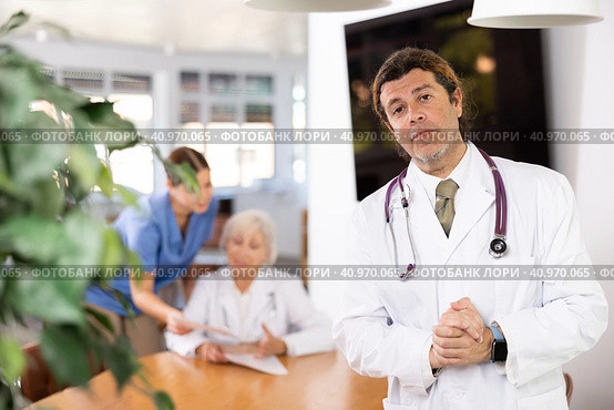 positive medical male colleague standing in meeting room and employees are talking behind him at table. Стоковое фото, фотограф Яков Филимонов / Фотобанк Лори