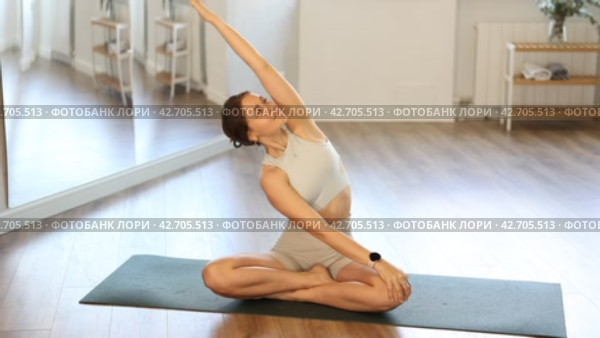 Positive young fit girl in sportswear doing yoga in modern mirrored studio, concentrating on performing revolved easy pose, basic seated posture with crossed legs and twist increasing flexibility. Стоковое видео, видеограф Яков Филимонов / Фотобанк Лори