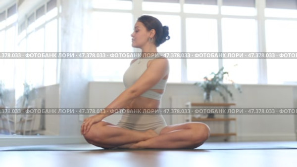 Positive young fit girl in sportswear doing yoga in modern mirrored studio, concentrating on performing revolved easy pose, basic seated posture with crossed legs and twist increasing flexibility. Стоковое видео, видеограф Яков Филимонов / Фотобанк Лори
