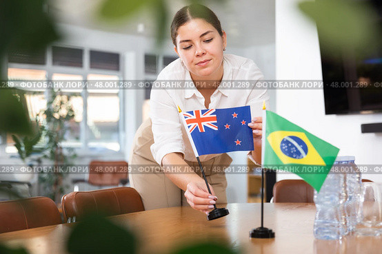 Positive young woman putting flag of Australia on the table with flag of Brazil in conference room. Стоковое фото, фотограф Яков Филимонов / Фотобанк Лори