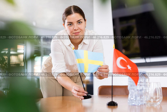 Positive young woman putting flag of Sweden on the table with flag of Turkey in conference room. Стоковое фото, фотограф Яков Филимонов / Фотобанк Лори