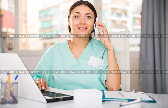 Practicioner woman in medical uniform talking by phone at laptop. Стоковое фото, фотограф Яков Филимонов / Фотобанк Лори