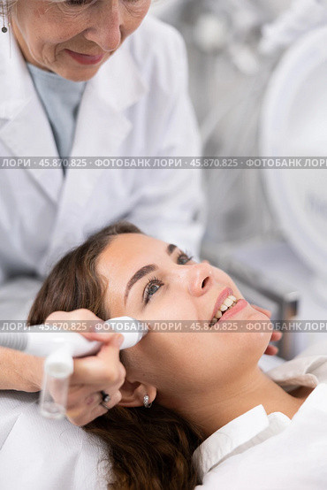 Professional mature female cosmetologist doing needleless high frequency mesotherapy in spa center. Non-invasive skin rejuvenation procedure. Стоковое фото, фотограф Яков Филимонов / Фотобанк Лори
