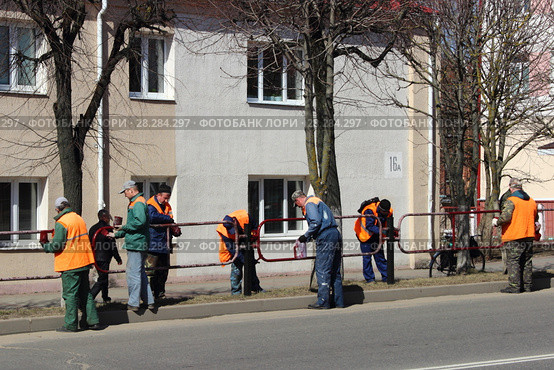 Рабочие маляры в спецодежде ЖКХ весной красят забор, дорожное ограждение на фоне дома и дороги (2018 год). Редакционное фото, фотограф Илья Илмарин / Фотобанк Лори