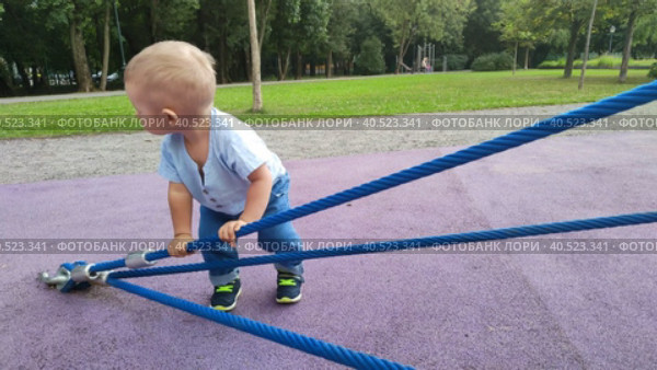 Ребенок играет в парке или во дворе летним днем. Игровая площадка детского сада. Маленький мальчик тянет толстые канаты на веревочной горке. Малыш гуляет на улице. Первые шаги. Дети познают мир. The child plays in the park or in the yard on a summer day. Kindergarten playground. A little boy pulls thick ropes on a rope slide. The kid is walking on the street. The first steps. Children learn the world. Стоковое видео, видеограф Ирина / Фотобанк Лори