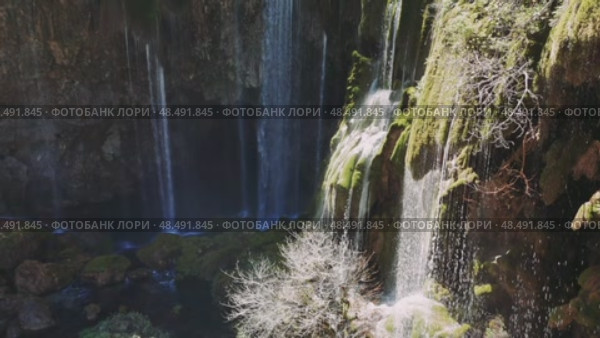 Rocky landscape with verdant moss, cascading waterfall streaming down lush terrain under bright sunlight, creating tranquil wilderness scenery. Стоковое видео, видеограф ILIA GOROKHOV / Фотобанк Лори