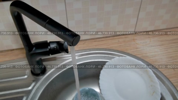 Рука открывает водопроводный стильный черный кран на кухне. Вода течет в раковину с грязной посудой. Крупный план. Оригинальный звук. Редакционное видео, видеограф Sergei Gorin / Фотобанк Лори