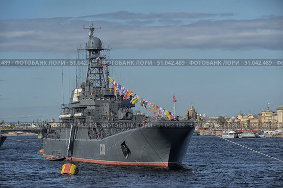 Russian Navy warship No. 130 is anchored in the Neva River in St. Petersburg. The vessel is decorated with flag colors of signal flags stretching from the mast to the bow and stern, which gives it a festive and solemn look. The Russian naval ensign flies from the mast, and the St. Andrew's flag flies from the stern. In the background, the historic buildings and golden domes of St. Petersburg's churches are visible, creating a contrast between the modern warship and the architectural heritage of the city. Clear weather, blue skies and calm water emphasize the atmosphere of a solemn maritime event, probably related to a parade or a Navy holiday. (2015 год). Редакционное фото, фотограф Анна Кривицкая / Фотобанк Лори