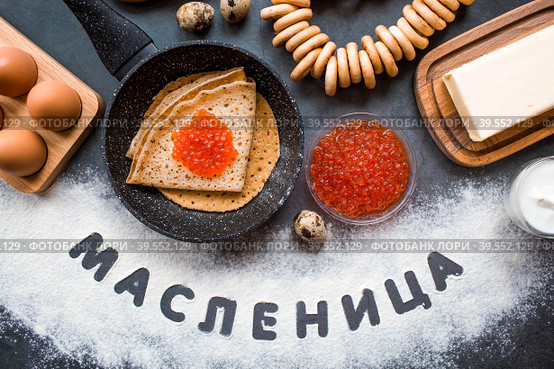 Русские традиции. Русский праздник Масленица. Стопка блинчиков, красная икра, бублики, рогалики, деревянная ложка, яйца и сливочное масло. Надпись на русском языке "Масленица" написано мукой. Вид сверху. Стоковое фото, фотограф Photojuli / Фотобанк Лори