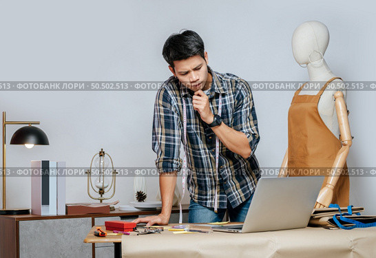 Seriously young Asian fashion designer or tailor working with laptop computer to search idea or online orders, full of tailoring tools with mannequin and equipment on the desk in the studio. Стоковое фото, агентство Ingram Publishing / Фотобанк Лори