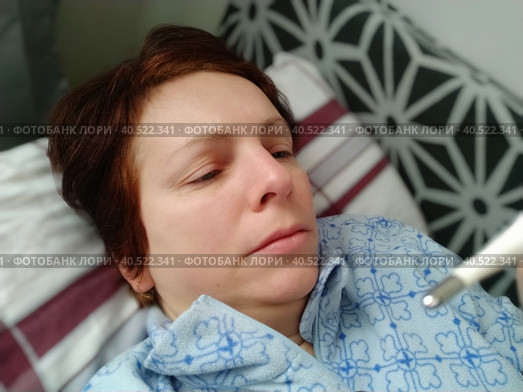 Sick woman in bed. Headache, runny nose and cold. Paleness of the face and faintness of the body. Feeling unwell. The patient lies on a pillow, holds a thermometer and measures the temperature. Стоковое фото, фотограф Юлия Петровская / Фотобанк Лори