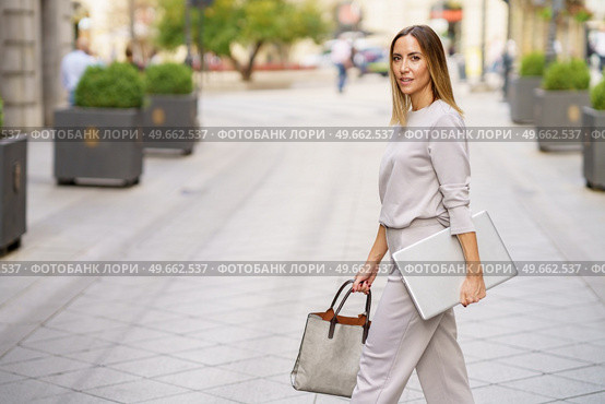 Side view of confident adult female manager in stylish outfit with blond hair looking at camera, while walking on paved street with laptop and bag in hands. Stylish female entrepreneur with netbook strolling on city street. Стоковое фото, агентство Ingram Publishing / Фотобанк Лори
