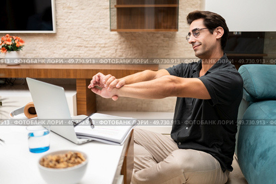 Side view of young male freelancer in casual clothes sitting near laptop and notebook while stretching arms at home. Happy young man stretching arms near netbook at home. Стоковое фото, агентство Ingram Publishing / Фотобанк Лори