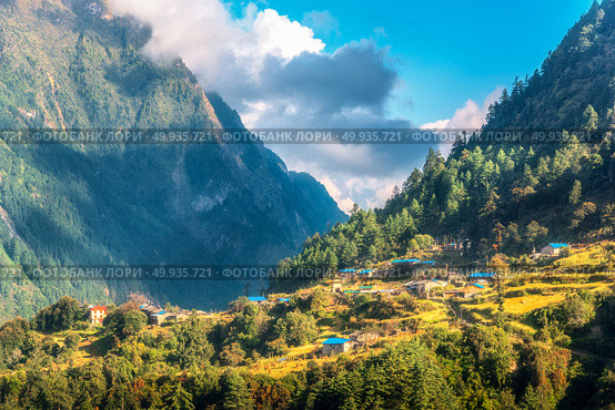 Small village on the hill lighted by a sunbeam against mountains and blue sky with clouds at sunset. Summer landscape with houses, fields with grass, gardens, green trees in Nepal. Rustic view. Travel. Small village on the hill lighted by a sunbeam against mountains. Стоковое фото, агентство Ingram Publishing / Фотобанк Лори
