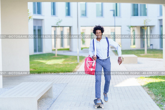 Smiling African American male entrepreneur with handbag riding skateboard along street in summer and looking at camera while commuting to work. Positive black businessman riding skateboard in city. Стоковое фото, агентство Ingram Publishing / Фотобанк Лори