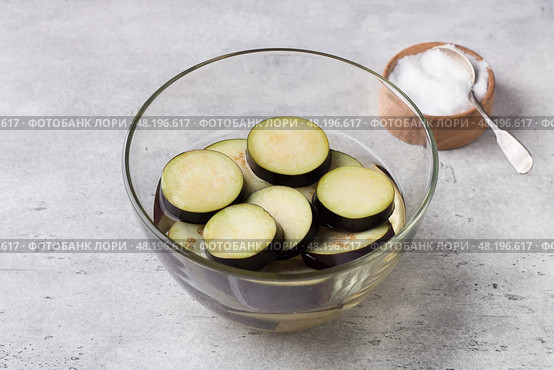 Soaking eggplant slices in salt water in a glass bowl on a gray textured background. Processing eggplant before cooking, removing bitterness. Стоковое фото, фотограф Ольга Лепёшкина / Фотобанк Лори