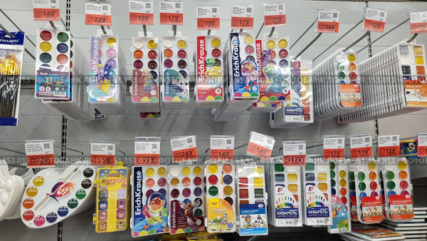 Store shelves with a wide range of watercolor paints. The display case features sets from different brands, including Erich Krause, Luch, Gamma and other manufacturers. The palettes are varied: classic round and rectangular trays, watercolors with pastel shades, as well as paints in original packages with cartoon characters. The number of colors varies from 6 to 18, there are sets with brushes and a palette. Each package has a bright design, aimed at both children and schoolchildren and aspiring artists. Above the products are price tags with promotional discounts. The image shows neatly laid out drawing supplies and is perfect for illustrating the range of stationery stores, online catalogs, educational projects and advertising materials. Редакционное фото, фотограф Анна Кривицкая / Фотобанк Лори