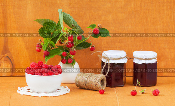 Summer rural still life with ripe freshly picked raspberries in bowl, jars of homemade jam and bouquet of raspberry branches with berries on wooden garden table on July day. Harvesting organic  berries. Стоковое фото, фотограф Виктория Катьянова / Фотобанк Лори