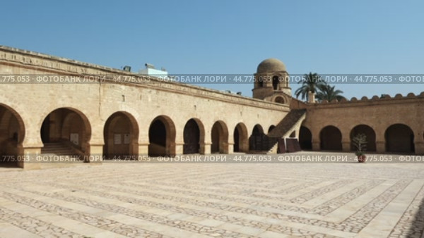 Sunlit paved courtyard in Great Mosque of Sousse surrounded by arched galleries with Kufic inscription band on walls, stone stairs to roof and domed corner tower under blue Tunisian sky. Стоковое видео, видеограф Яков Филимонов / Фотобанк Лори