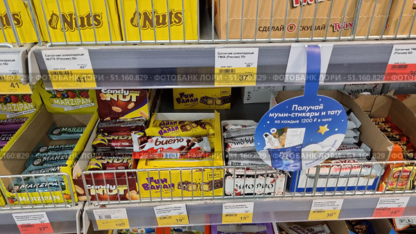 Supermarket display cases with chocolate bars and sweets from various brands. In the foreground, you can see brightly colored packages of Nuts, Twix, Kinder Bueno, Fun Banan, Candy Nut, Marzipan and other bars. All the products are neatly arranged in boxes with price tags indicating the cost and weight. The display case is made in a standard self-service store, with metal shelves and dividers. In the center of the photo, there is an advertising sign offering Moomin stickers and tattoos for purchases over a certain amount. The bright composition emphasizes the variety of products and the atmosphere of a grocery store. (2021 год). Редакционное фото, фотограф Анна Кривицкая / Фотобанк Лори