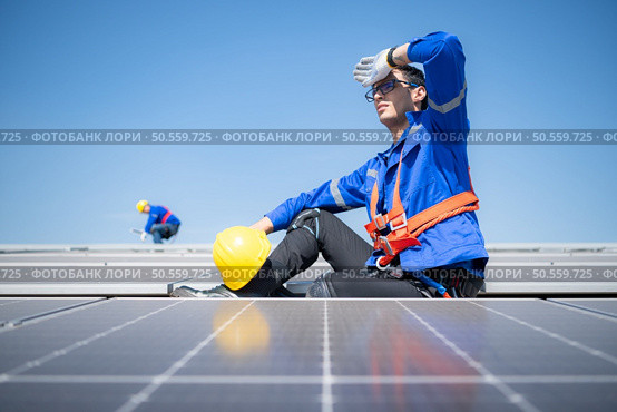 Technician repairing solar panels take off his hat and rest in the scorching sun on a factory roof covered with solar panels to receive solar energy. Стоковое фото, агентство Ingram Publishing / Фотобанк Лори