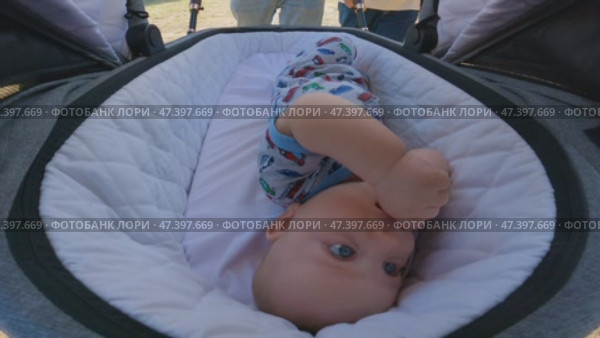 The baby is lying in stroller, dressed in a colorful jumpsuit with pictures of cars. The stroller is covered with white quilted fabric. The parents push stroller through park. A close-up of baby. Стоковое видео, видеограф Воловодов Владимир / Фотобанк Лори