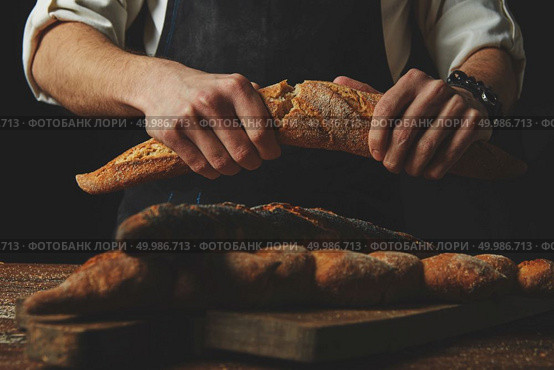 The baker’s hands break a delicious fresh crispy baguette on a dark wooden background. Baker’s hands break the baguette. Стоковое фото, агентство Ingram Publishing / Фотобанк Лори