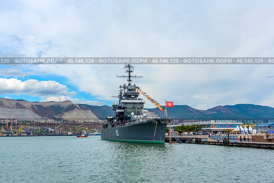 The cruiser Mikhail Kutuzov, permanently moored at the Novorossiysk embankment. This is one of the main museum ships of Russia, which is part of the Central Naval Museum.The cruiser Mikhail Kutuzov is a massive warship with the number "105" on board.It is decorated with signal flags, creating a festive atmosphere.In the background, the port of Novorossiysk, the mountains of the Markotkhsky Range and the berthing facilities are visible.The red naval flag with a white star flutters at the stern.To the right of the ship is the marine terminal, where people and city infrastructure are visible.In the distance, a small tug or boat moves along the water. (2023 год). Редакционное фото, фотограф Анна Кривицкая / Фотобанк Лори