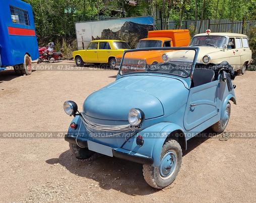 The legendary Soviet micromobile SMZ S-3D, also known as the "invalidka". This is a lightweight open-top vehicle designed for people with disabilities, especially war veterans. The car is a two-seater, with a simplified design: rounded shapes, small wheels, headlights protruding from the fenders, a soft awning rolled up and secured at the back. The car is painted matte blue. The SMZ S-3D was produced at the Serpukhov Motorcycle Plant and became a symbol of accessible transport in the Soviet era.The car is on display at a retro-car exhibition among other classic cars: a Volga in the back of an ambulance, a Moskvich, a van and a bright Zhiguli are visible nearby. The atmosphere is like an open-air museum. Стоковое фото, фотограф Анна Кривицкая / Фотобанк Лори