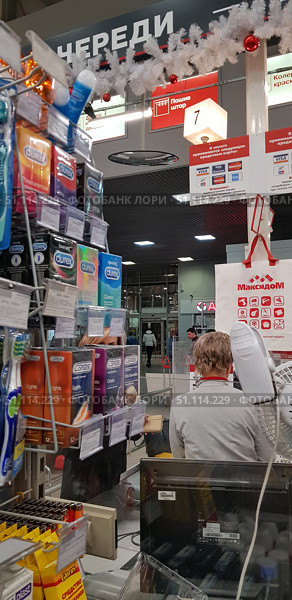 The photo was taken in a checkout line at a large shopping center. The frame shows the checkout area with a salesperson, customers, and a counter with impulse goods. The display case contains packages of condoms from well-known brands Durex and Contex, as well as related small goods - toothbrushes, batteries, patches. In the background, the sign of the Maksidom store and the checkout number 7 are visible. Above the checkouts, there is a festive decoration - a white garland with red balls. The information signs indicate payment methods, including Visa and MasterCard bank cards. The atmosphere of everyday shopping and excitement in a department store. (2018 год). Редакционное фото, фотограф Анна Кривицкая / Фотобанк Лори