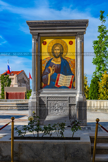 The photograph shows a close-up of the icon of Jesus Christ installed in a massive carved stone icon case on the territory of the New Chersonesus cultural and historical complex in Sevastopol. The icon is made in the traditional Orthodox Byzantine style. In the background are visible architectural elements in the Neo-Byzantine style, the Russian flag, decorative trees and a blue sky with light clouds. The atmosphere of the frame is filled with solemnity and spiritual significance. Стоковое фото, фотограф Анна Кривицкая / Фотобанк Лори