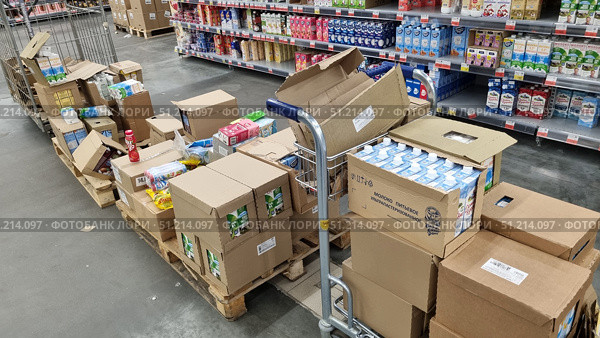 The process of displaying goods in a supermarket. In the foreground are wooden pallets with cardboard boxes, some of which are open. The boxes contain packages of milk, yogurt and other dairy products. A trolley for moving boxes is visible. In the background is a retail shelving unit with a large assortment of dairy products: milk, cream, kefir, yogurts in cardboard packages of different brands. The atmosphere of the photo conveys the work process of the store, the organization of supplies and logistics. Редакционное фото, фотограф Анна Кривицкая / Фотобанк Лори