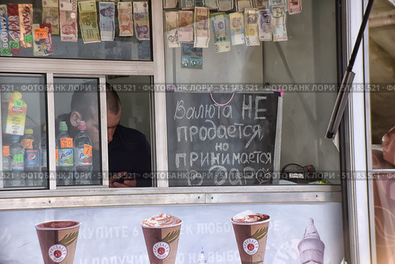 The window of a street kiosk or small shop. In the foreground hangs a chalk board with the inscription: "Currency is not sold, but accepted as a gift." Above the window are various banknotes from different countries of the world, used as a decorative element. Inside the kiosk, behind the glass, you can see bottles of water and fizzy drinks with price tags, as well as chocolate bars and juices. Behind the counter is a seller, intently looking at his phone. Below is part of an advertising banner with an image of coffee and ice cream. The atmosphere of the photo combines humor, everyday life and street trading. (2015 год). Редакционное фото, фотограф Анна Кривицкая / Фотобанк Лори
