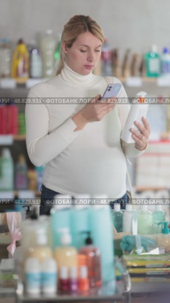 Thoughtful pregnant woman scanning bottle of sunscreen in personal care store using phone, checking ingredients to find safest option for her and future baby before making mobile payment. Стоковое видео, видеограф Яков Филимонов / Фотобанк Лори