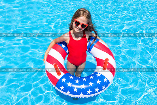Top view happy little girl in red swimsuit and sunglasses stands in outdoor swimming pool with a lifebuoy decorated with American flag print. Vacation in USA concept. 4th of July pool party. Стоковое фото, фотограф Ирина Михайличенко / Фотобанк Лори