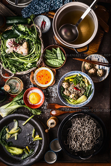 Top view of Asian food dishes. Asian noodle soup preparation on dark rustic kitchen table. Pot with stock, cooked noodles, various vegetables and meatballs. Meatball noodle soup in bowl with chopstick. Стоковое фото, агентство Ingram Publishing / Фотобанк Лори