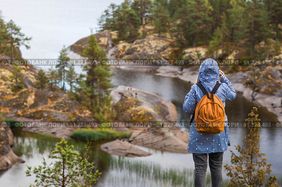 Tourism. Hiking. Ladoga skerries on Lake Ladoga. A female tourist in a raincoat with a backpack on the rocky islands looks into the distance. Republic of Karelia, Russia. Стоковое фото, фотограф Photojuli / Фотобанк Лори