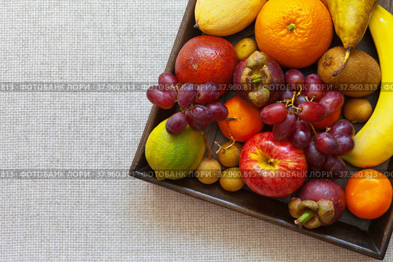 Tropical ripe juicy fruits on a wooden tray on a table with a linen tablecloth. Banana, orange, mango, grapes, tangerines, mangosteen, lychee, apple, pear, kiwi, lime. Healthy diet. Top view, flat lay, copy space. Стоковое фото, фотограф Виктория Катьянова / Фотобанк Лори